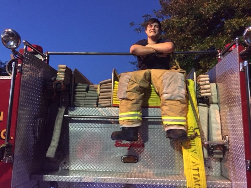 A young firefighter perched on top of the engine's hosebed at dusk